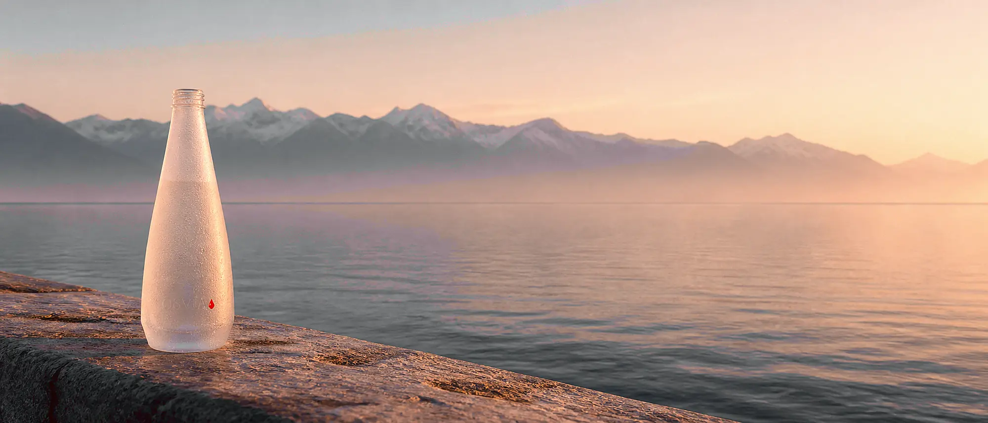 Au bord du Léman à l'aube, la bouteille OSVIA sablée posée sur un parapet de pierre, les Alpes en silhouette dans la brume